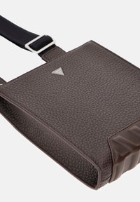 Ciemnobrązowa torebka na ramię z teksturowanej skóry z czarnym regulowanym paskiem i małym srebrnym trójkątnym logo z przodu.