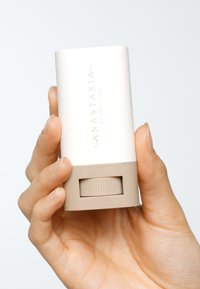 Envase cilíndrico blanco para cuidado de la piel con base beige, mecanismo de giro texturizado y letras doradas para "Anastasia Beverly Hills."