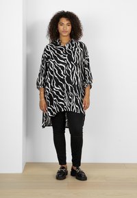 Chemise longue en imprimé zèbre noir et blanc avec des manches 3/4, ourlet droit, associée à un pantalon noir et des chaussures noires brillantes.