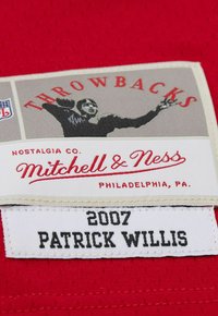 Maillot de sport en tissu rouge avec un motif texturé. Présente une étiquette grise avec "THROWBACKS", et un graphique d'un joueur, marqué "Mitchell & Ness."