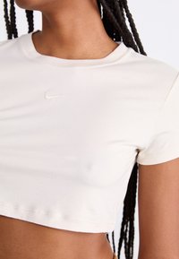 Šviesiai smėlinės spalvos trumpa palaidinė, pagaminta iš minkštos medžiagos, su apvalia iškirpte ir mažuoju išsiuvinėtu Nike logotipu ant krūtinės. Trumpomis rankovėmis.