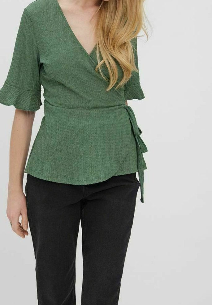 Femme portant un chemisier vert cache-cœur avec des coudes volantés et un pantalon noir, debout devant un fond clair uni.