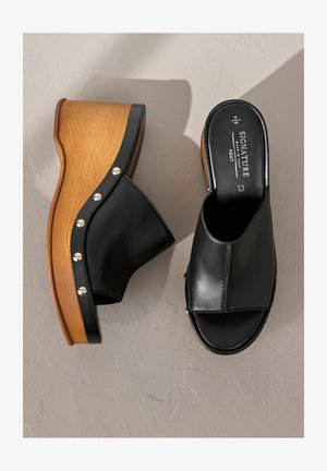 Czarne skórzane sandały na koturnie z platformą przypominającą drewno, srebrnymi ćwiekami wzdłuż krawędzi i otwartym noskiem. Teksturowana podeszwa zapewnia przyczepność.