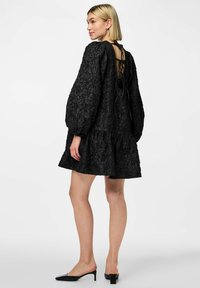 Vestido negro texturizado con falda acampanada, mangas abullonadas y espalda descubierta con un detalle de cordones. Tejido suave y acentos en contraste.