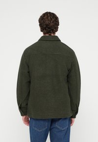 Vihreä villasekoitteinen takki, jossa on kaulus, pitkät hihat ja näkyvät ompelusaumat. Yhdistetty sinisiin farkkuihin, jotka ovat rentoa mallia.