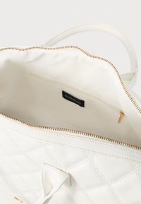 Λευκή τσάντα χειρός με καπιτονέ σχέδιο, χρυστό φερμουάρ και εσωτερική τσέπη. Λείο υλικό με απαλή υφή και εσωτερική ετικέτα με λογότυπο.