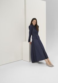 Abito in denim scuro con giacca su misura e gonna fluente. Tessuto liscio, maniche lunghe e corpetto aderente. Scarpe tacco grigio chiaro completano il look.