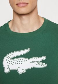 Vihreä puuvillainen t-paita, jossa on suuri valkoinen krokotiilikuva kolmio-kuvioilla, pyöreä pääntie ja lyhyet hihat.