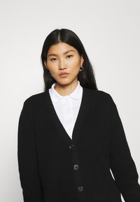 Schwarzer gerippter Cardigan mit V-Ausschnitt und sechs Knöpfen. Über einem weißen Hemd mit Kragen getragen, hat er ein strukturiertes Material und lange Ärmel.