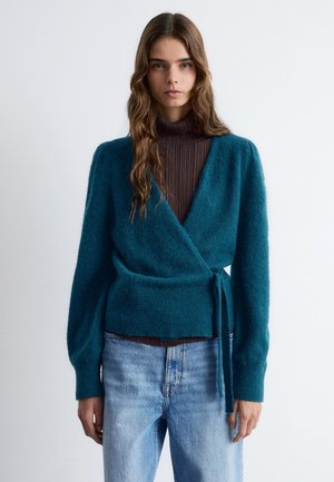 Teal wrap cardigan made from a soft, fuzzy material. Features long sleeves, a tie at the waist, and a deep V-neckline. Worn over a brown turtleneck.