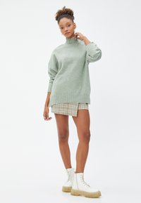 Jasnozielony sweter dziany z wysokim kołnierzem i objętościowymi rękawami, w połączeniu z spódnicą w kratę i białymi masywnymi butami. Minimalistyczny design.