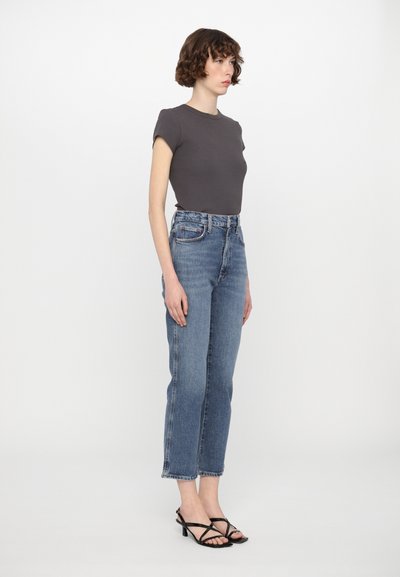 Grå åtsittande t-shirt, hög midja ljusblå denimjeans, svarta sandaler med klack och remmar. Modellen står i profil mot en vit bakgrund.