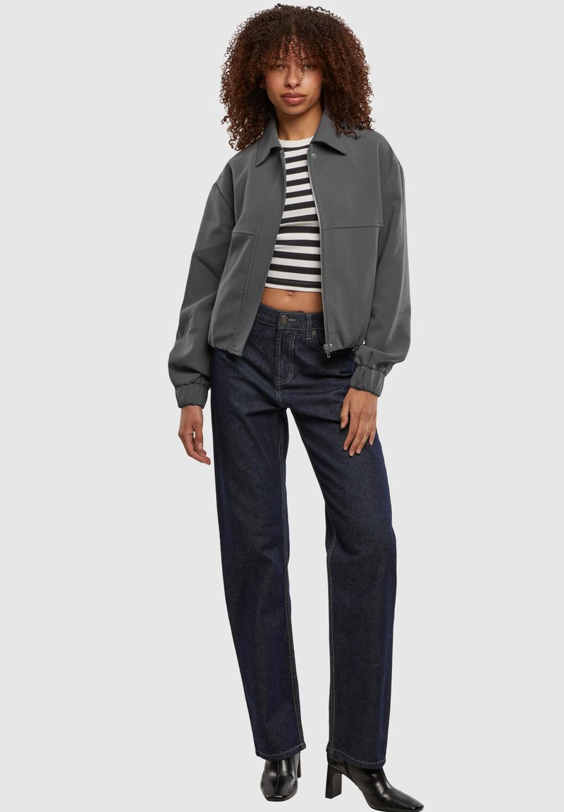 Frau mit lockigen Haaren, die eine graue Reißverschlussjacke, ein schwarz-weiß gestreiftes kurzes Top, dunkle Jeans und schwarze Stiefel mit Absatz trägt.