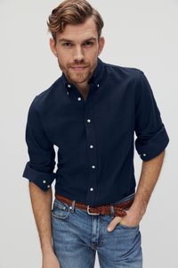 Navyblauwe button-up shirt van katoen met opgerolde mouwen, gecombineerd met lichtblauwe jeans en een gevlochten brown leren riem, model recht vooruit kijkend.
