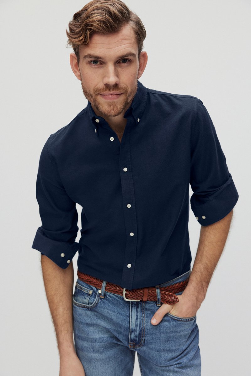 Navyblauwe button-up shirt van katoen met opgerolde mouwen, gecombineerd met lichtblauwe jeans en een gevlochten brown leren riem, model recht vooruit kijkend.