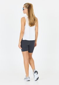 Top bianco senza maniche, pantaloni corti scuri e scarpe da ginnastica grigie. Capelli lunghi, occhiali da sole blu, che si allontana dalla macchina fotografica.