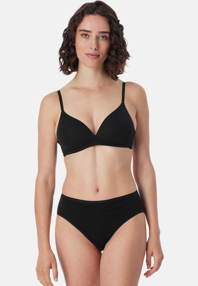 Frau mit lockigem Haar, die einen schwarzen BH und passende schwarze hochtaillierte Unterwäsche trägt, steht vor einem einfachen weißen Hintergrund.