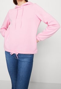 Felpa rosa pallido in vestibilità rilassata, con cappuccio regolabile e polsini elastici. Abbinata a jeans blu. Texture morbida, design minimale.