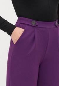 Vijolične hlače imajo visok pas z naborki, stranske žepke in dva črna gumba spredaj, narejene so iz teksturiranega materiala.