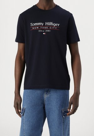 Mężczyzna w granatowej koszulce Tommy Hilfiger z napisem "New York City Since 1985" oraz jasnoniebieskich spodniach typu cargo z denimu.