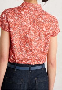 Blusa corallo a maniche corte con motivo floreale bianco, colletto con bottoni e vestibilità rilassata, abbinata a jeans scuri e una cintura blu.