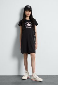 Juodas medvilninis marškinėlių suknelė su trumpomis rankovėmis, kurios priekyje yra rožinė grafika. Dėvima su baltomis sportinėmis batelėmis ir juoda kepure.