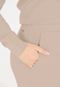 Beige joggers met een elastische tailleband en zijzakken, met een subtiele glans en een gouden merkteken op de tailleband.