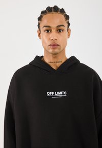 Ungdom med flettehår iført svart hettegenser med teksten "FORBUDT, FARLIG OMRÅDE," som viser en nakketatovering med teksten "tro på deg selv."