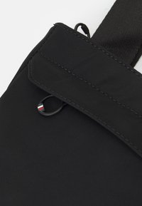 Borsa nera in nylon con patta frontale con dettagli cuciti, un accento con anello in metallo e una superficie testurizzata. Cinghia regolabile inclusa.