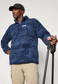 Navyblauwe fleece pullover met een gestructureerd patroon, kwart ritssluiting en borstzak; gedragen met khaki broeken en met stokken in de hand.