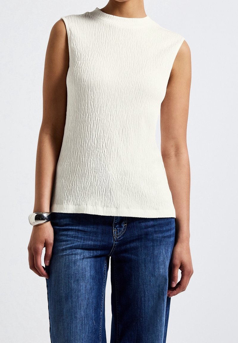 Haut blanc texturé sans manches assorti à un jean bleu et un bracelet argenté au poignet gauche, porté par une personne debout.