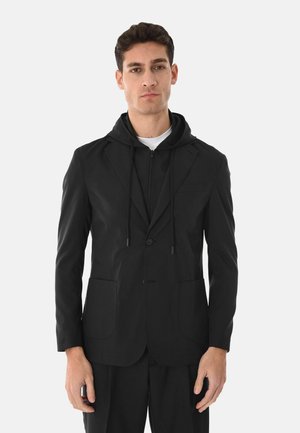 Uomo che indossa un blazer nero con cappuccio incorporato sopra una camicia bianca, in piedi contro uno sfondo chiaro uniforme.
