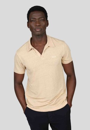 Polo beige à manches courtes et col, présentant une texture subtile et un petit logo blanc sur la poitrine.