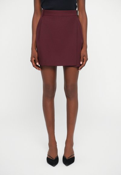 Mini-jupe bordeaux avec une texture lisse, taille mi-haute et plis sur les côtés, associée à des escarpins noirs à bout pointu.