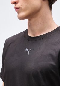 Κοντινό πλάνο ενός ατόμου που φοράει ένα απλό μαύρο t-shirt Puma, δείχνοντας το πάνω μέρος του στήθους και το πλαϊνό προφίλ του λαιμού και του προσώπου.