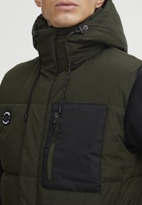 Grüne Pufferweste mit hohem Kragen, schwarzem Brusttaschen, Kordelzugkapuze und Reißverschlussdetails. Weicher Stoff und gestepptes Design.