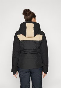 Svart og beige boblejakke med hette, med horisontal søm, polstret tekstur og røde aksenter på skuldrene. Vist fra bak.
