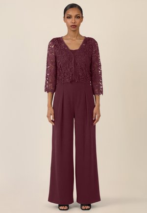 Frau steht, trägt eine bordeauxrote Spitzenstrickjacke mit Dreiviertelärmeln über einem passenden ärmellosen Overall und schwarzen offenen Schuhen mit Absatz.