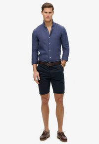 Navyblauwe overhemd met lange mouwen en knopen, donkergekleurde shorts, bruine leren riem en boot schoenen. Linnen structuur, geen patronen en slim fit ontwerp.
