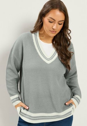 Kvinde med langt brunt hår iført en løs grå strikket sweater med hvid stribet kant, står med hænderne i lommerne og kigger ned.