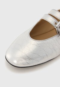 Chaussure plate en cuir argenté avec un bout pointu, présentant une texture craquelée et deux brides réglables avec un petit détail de boucle.