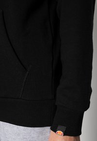 Sweat-shirt noir avec poignets et ourlet côtelés, doté d'une poche kangourou et d'une étiquette logo sur la manche. Le tissu semble doux et texturé.