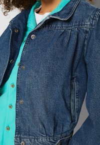 Mörkblå denimjacka med knäppning fram, krage, elastisk midja och sidofickor; lagd över en ljus turkos skjorta.