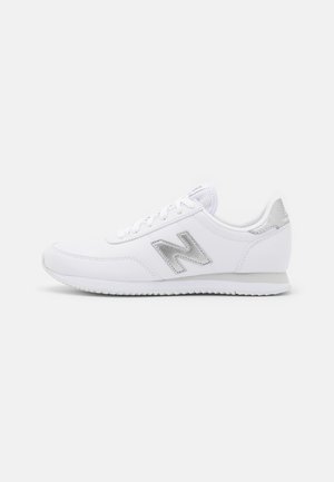 Zapatilla blanca New Balance con logo "N" gris, diseño con cordones y suela de goma de perfil bajo sobre un fondo claro liso.