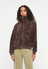 Bruine fleece jas met hoge kraag, ritssluiting en voorzakken. Beschikt over een zachte textuur en ribgebreide manchetten en zoom. Gemaakt om te dragen met licht corduroy broeken.