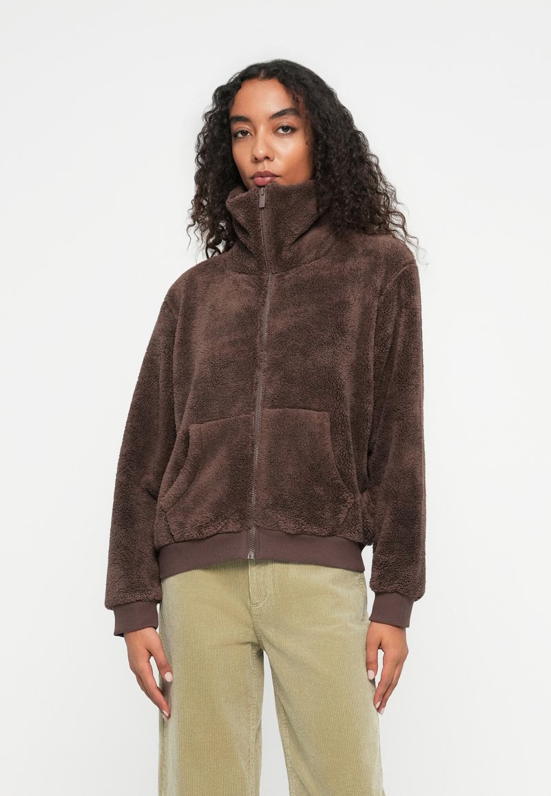 Bruine fleece jas met hoge kraag, ritssluiting en voorzakken. Beschikt over een zachte textuur en ribgebreide manchetten en zoom. Gemaakt om te dragen met licht corduroy broeken.