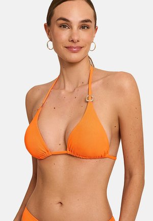 Femme portant un haut de bikini orange à col halter avec des détails de boucle dorée et des boucles d'oreilles anneaux, souriante sur un fond uni.