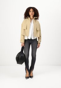 Veste beige courte avec grandes poches, chemise blanche à boutons, jeans foncés avec fentes sur le côté, et sac noir à la texture lisse et à la forme arrondie.