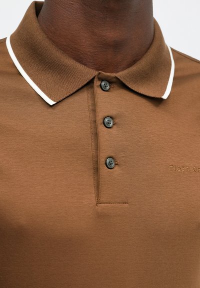 Hnědá polo košile z hladkého materiálu, s bílým lemem límce, třemi černými knoflíky a vyraženým logem na hrudi.
