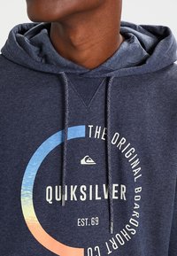 Sudadera con capucha de forro polar azul marino con cordones, con un logo gráfico circular en tonos azul y naranja, y un acabado texturizado.